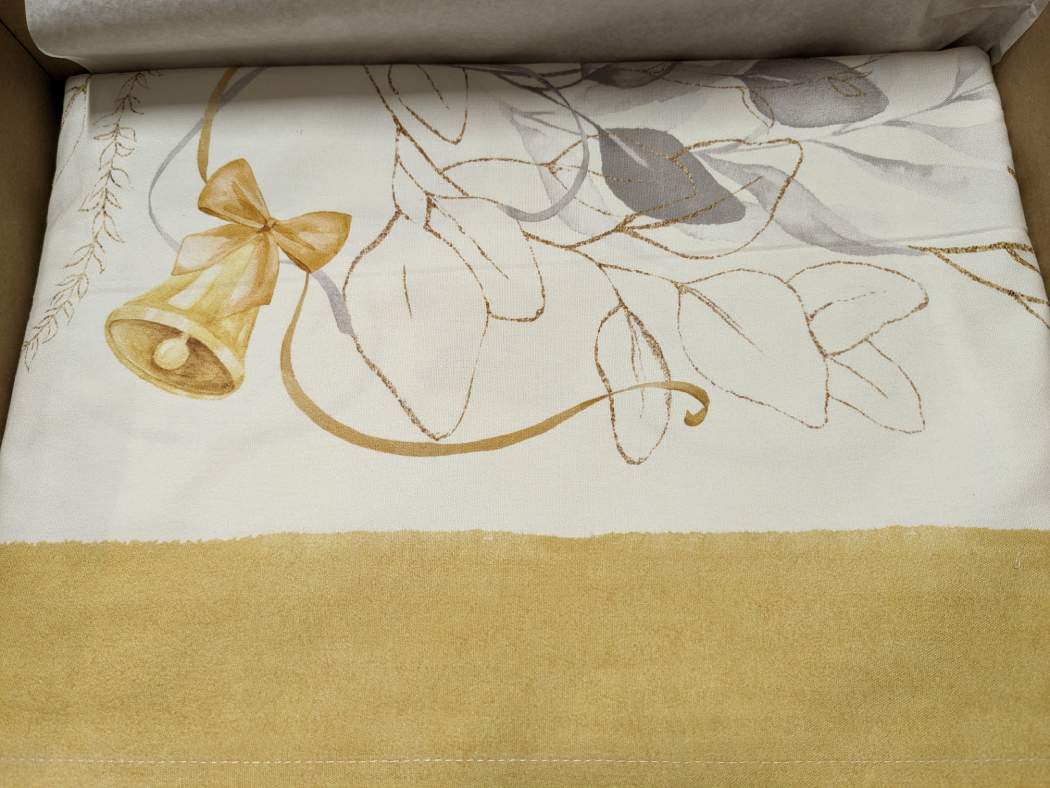 Tovaglia natalizia Mirage di Tessitura Randi. Stampa digitale su panama di puro cotone. Fantasia su fondo avorio con delicata fantasia con foglie, nastrini ed elementi tipici del Natale nei colori oro e grigio. Disponibile nella misura rettangolare x12 (150x270 cm) o x18 (150x360 cm). dettaglio fantasia bordo ocra