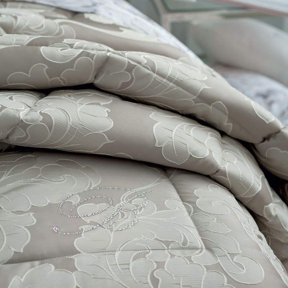 Trapunta Invernale matrimoniale Gemma di Blumarine, in elegante raso jacquard.Fantasia tinta in filo dalle forme morbide, nella variante di colore tortora, con logo Blumarine in preziosi cristalli Swarovski. Dettaglio fantasia.