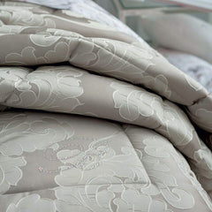 Trapunta Invernale matrimoniale Gemma di Blumarine, in elegante raso jacquard.Fantasia tinta in filo dalle forme morbide, nella variante di colore tortora, con logo Blumarine in preziosi cristalli Swarovski. Dettaglio fantasia.