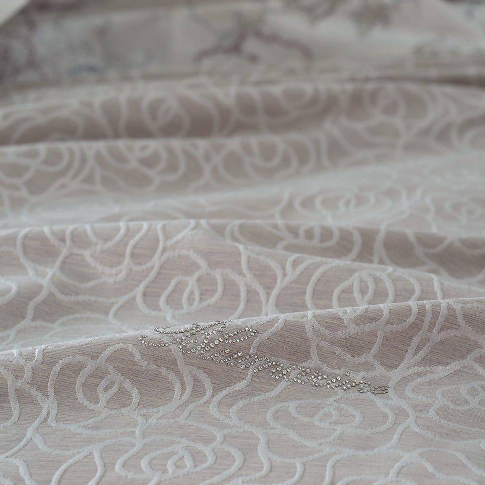 Copriletto estivo matrimoniale Leslie di Blumarine, in elegante raso jacquard. Romantica fantasia jacquard con linee sinuose che si intrecciano a formare delle rose, colore beige/tortora, con logo Blumarine in preziosi cristalli Swarovski. Coperta estiva