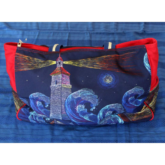 In spiaggia con stile, con la borsa da mare Faro di Karakorum.  La borsa mare presenta una fantasia con faro e onde su fondo blu, con inserti rossi sui lati. Chiusura con zip e manici bicolore (blu e rossi). Tasca interna con zip